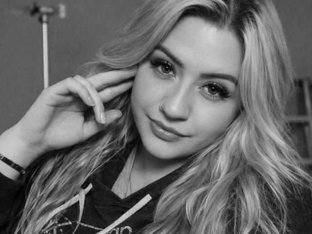 Black and white selfie of Arynata with long, wavy blonde hair, nose ring, and subtle makeup, wearing a hoodie, hand near ear, slightly smiling, looking at camera.