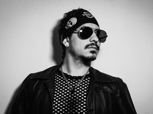 DavidBlake with a mustache, wearing a black bandana with peace signs, round sunglasses, polka dot shirt, leather jacket, and chain, looking to the side in a black-and-white shot.