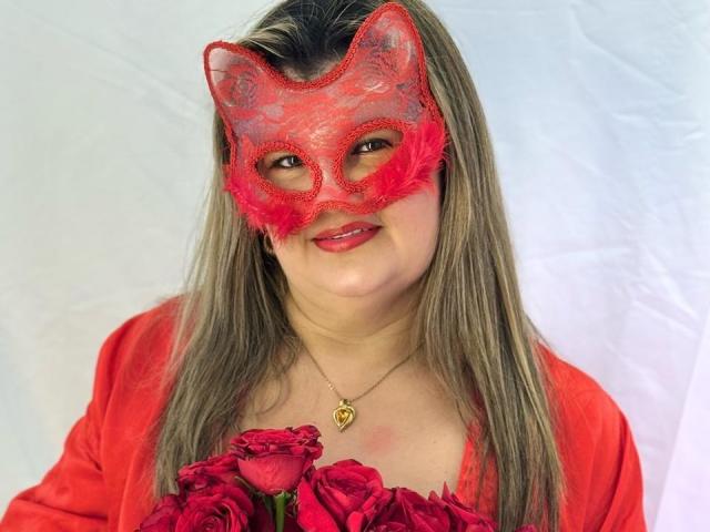 AdrianaPrettie with red lace mask, holding pink roses, wearing red shirt, gold necklace, and pink lipstick, smiling, long blonde hair, fair skin, pink mask with feather details.