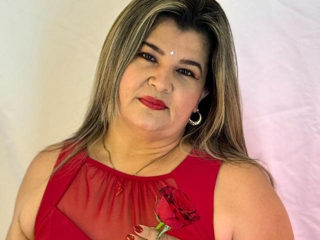 AdrianaPrettie has medium tits, a red dress with a rose, and a forehead bindi. She's smiling with red lipstick, wearing hoop earrings, and has blonde-streaked brown hair.