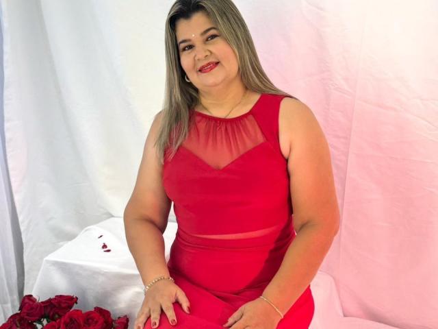 AdrianaPrettie with medium tits, curvy, in a red dress with sheer neckline, sitting, smiling, gray hair, red lipstick, white background, red roses, bracelet.