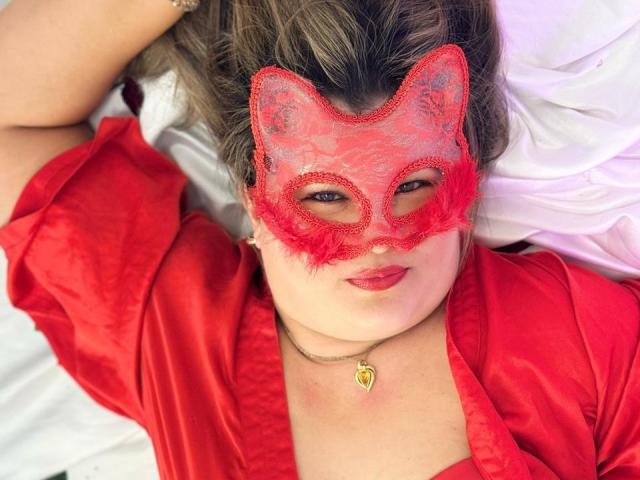 AdrianaPrettie with red lace mask, red dress, and heart necklace, lying on white sheets, smiling with red lipstick, brown wavy hair, and a cute, curvy, sexy look.