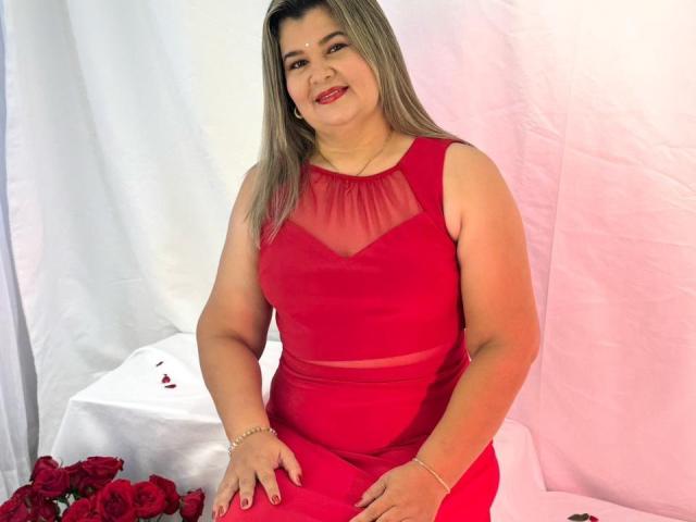 AdrianaPrettie has medium tits, a chubby ass, and is wearing a red dress with sheer shoulders. She's smiling with red lipstick, sitting among red roses.
