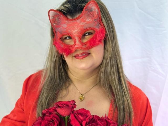 AdrianaPrettie wearing a red lace mask with pink feathers, holding red roses, has blonde hair, red shirt, and a gold heart necklace, smiling at the camera.