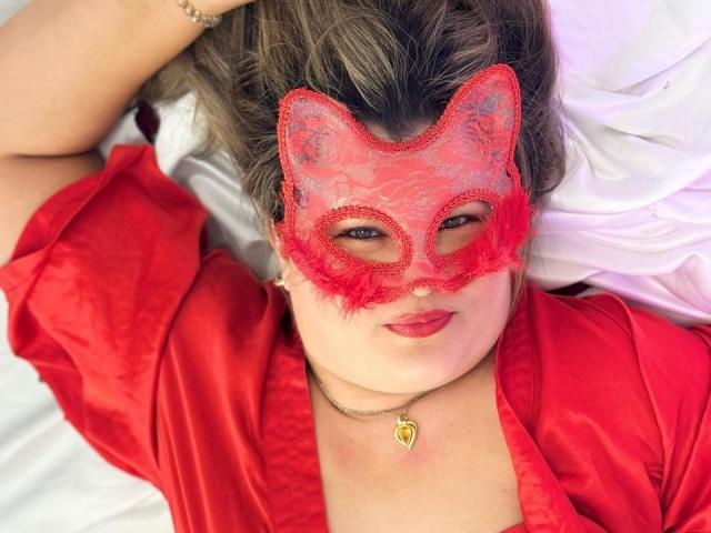 AdrianaPrettie in a red lace mask with feather edges, lying on white sheets, wearing a red satin robe, red lipstick, and a gold heart necklace. Her tits are partially visible.