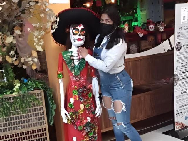 ArianithaXox stands next to a life-sized, painted, Day of the Dead figure with a red dress, green bow, and floral decorations. She wears a black mask, white jacket, and ripped jeans, touching the figure's chest. The figure has a black hat, white face with red markings, and green eyes. The scene includes a wooden shelf and green plants. ArianithaXox's right hand is on the figure's chest, left arm around its back. The image is a photograph.
