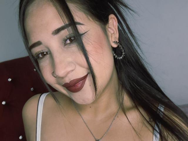 ViolletMoon con rossetto scuro, eyeliner nero, piercing al naso e orecchini a cerchio, sorride alla fotocamera, indossa una canotta bianca, mostrando il seno e una piccola lentiggine sul mento..