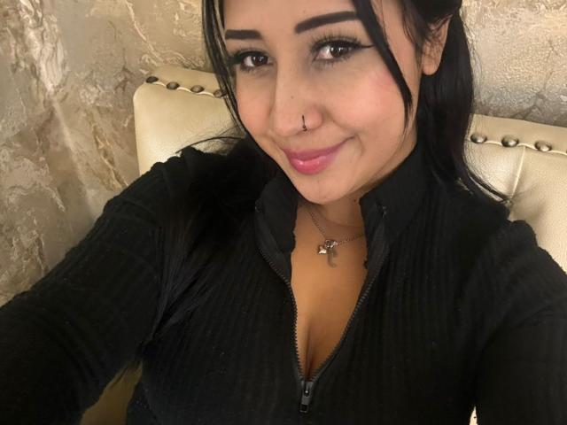 ViolletMoon, com cabelo escuro, piercing no nariz e batom rosa, vestindo uma jaqueta preta com zíper, mostrando os seios, sorrindo, sentada em uma cadeira bege com encosto cravejado..
