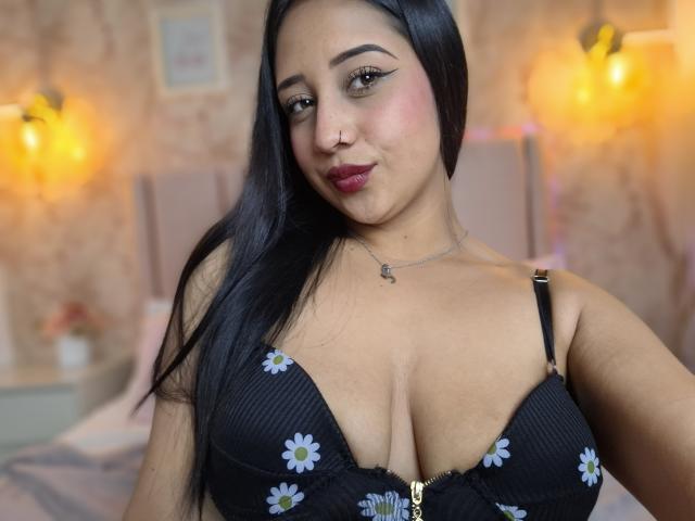 ViolletMoon, con lunghi capelli neri, indossa un reggiseno nero con fiori, mostrando il suo seno prosperoso, con un leggero trucco rosa sulle guance e un piercing al naso, sorride leggermente, in una stanza accogliente e illuminata da una luce calda..