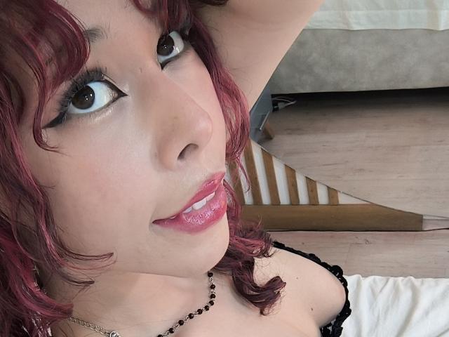SavanahReed has red curly hair, black eyeliner, pink lipstick, and is looking up with a slight smile. She's wearing a black necklace and a black top.