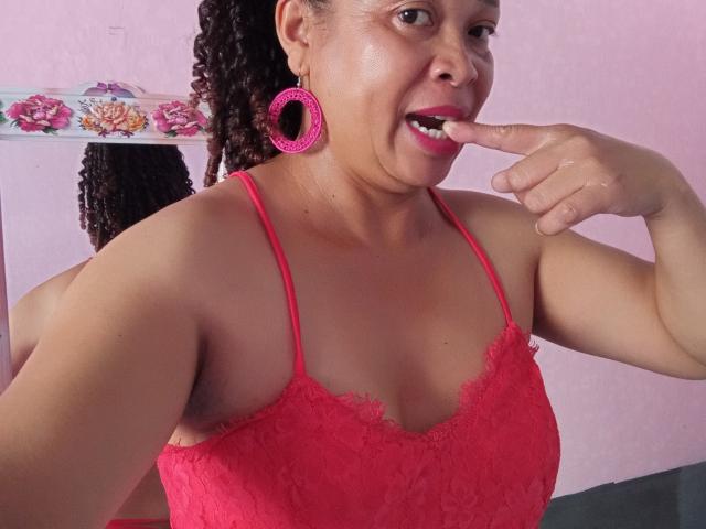 Rahel69 with curly brown hair, pink lace top, and matching pink hoop earrings, biting her finger with her lips slightly parted, showing off her small, firm tits.