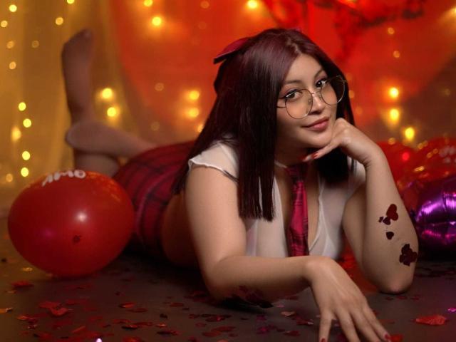 LeylaMiller, with glasses, red plaid skirt, and white top, lays on her stomach, legs up, showing her ass, red balloons, and butterfly tattoos on her arm.