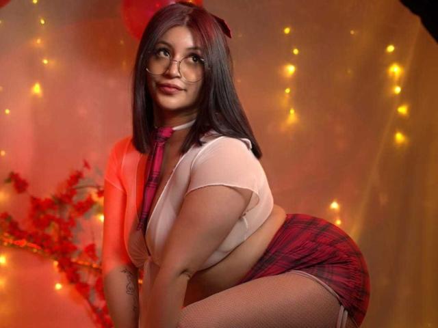LeylaMiller with dark brown hair, glasses, and a cute smile, wearing a red plaid skirt, white crop top, and pink tie, bent over, showing her big ass and small tits.