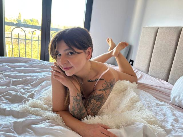 DaryX-hot laying on a bed, white sheets, arms tattooed, brown hair, pink bra, legs up, white furry blanket, smiling, windows in background, natural light, bare feet.