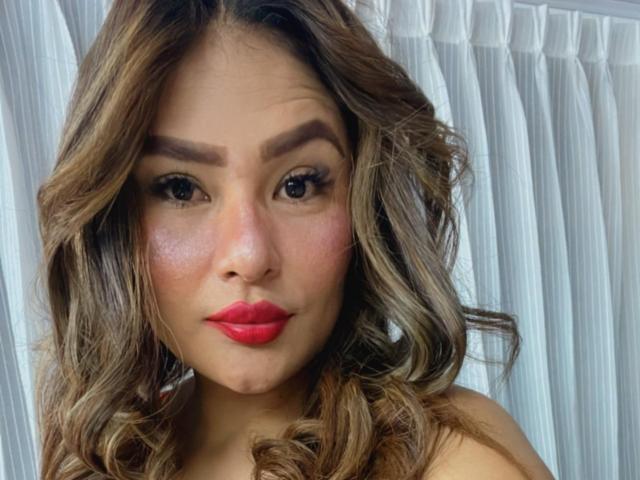 MiaaStar with wavy brown hair, red lipstick, thick eyebrows, and pinkish cheeks, looking directly at the camera with a subtle pout. Her tits are not visible.