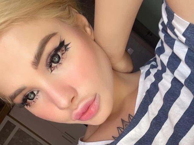 SweetNosha has blonde hair, green eyes with long eyelashes, pink lips, and a white and blue striped shirt, showing a tattoo on her neck. Her tits are partially visible.