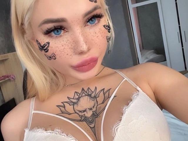Blonde SweetNosha with blue eyes, freckles, butterfly tattoos on cheeks, pink lips, white lace bra, and large tits, showcasing a lotus flower tattoo on her chest.