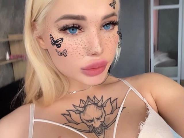 SweetNosha with blonde hair, blue eyes, and freckles, wearing a white lace top, showing a sunflower tattoo on her chest, and butterfly designs on her cheek.