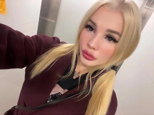 SweetNosha has blonde pigtails, thick eyelashes, and pink lipstick. She's wearing a maroon sweater and black straps, taking a selfie in a brightly lit elevator.
