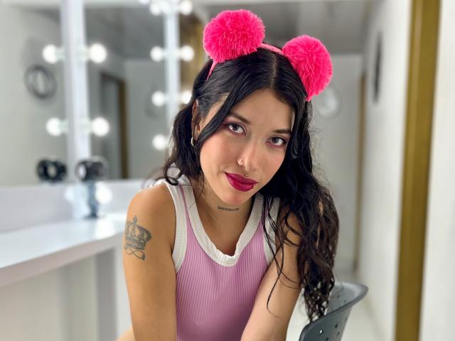 KrystalEvans with pink furry cat ears, pink ribbed tank top, long black hair, red lipstick, tattoo on left arm, sitting in front of mirror with lights.