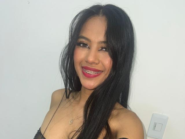 EstrellaXHot with long black hair, smiling with pink lipstick, showing her teeth, wearing a black strap dress, necklace with heart pendant, and looking cute.