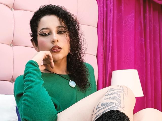 IrinaSweett with curly black hair, green top, heart necklace, tattooed thigh, pink headboard, and pink curtain, thinking with finger on chin, looking sexy.
