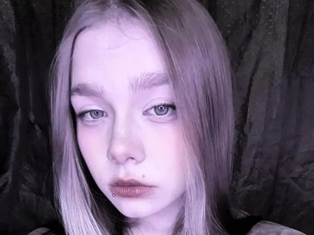 Lerinda has pale skin, straight platinum blonde hair, blue eyes, a small beauty mark on her left cheek, and a lower lip piercing. Her expression is serious and slightly sad.