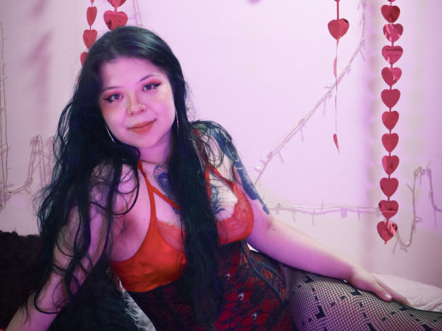 TrinityMoss with long black hair, red lace bra, and black fishnet stockings, sitting on a bed, wearing a red and black corset, smiling with pink eye makeup.