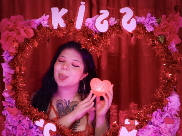 TrinityMoss with closed eyes, sticking out tongue, holding glittery red heart, wearing hoop earrings, orange dress, visible tattoo on chest, framed by red tinsel and purple flowers.