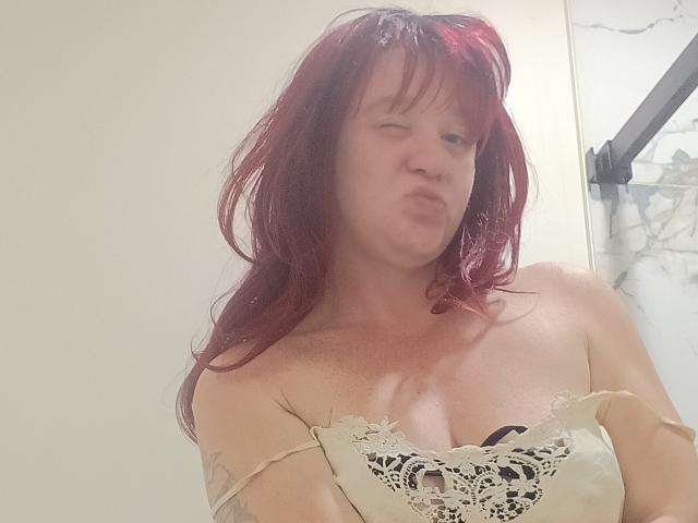 DeliciousRain with red hair, puffy lips, wearing a lacy off-shoulder top, exposing her tits, has a tattoo on her right arm, in a bathroom.