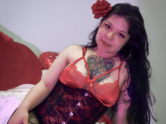 TrinityMoss with long black hair, red lace top, black corset, and rose hairpiece, showing a large tattoo on her chest, sitting on a bed with red pillows.