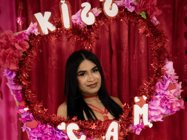 SilvanaCastro stands center, framed by a red glittery heart-shaped wreath with "KISS ME" letters. She has long black hair, dark makeup, and a red choker. Pink and purple paper flowers are attached to the wreath. The red curtain backdrop is simple, emphasizing the heart and SilvanaCastro's confident smile. The composition is straightforward, focusing on the subject and the decorative heart.