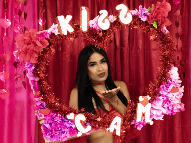 SilvanaCastro stands in front of a red curtain, framed by a heart-shaped garland of pink and red flowers with "KIS ME" in glowing neon letters. She has long black hair, dark eyebrows, and is wearing a red choker and matching red lingerie. She holds a red ribbon in her right hand, with her lips slightly puckered in a kissy face. The garland and neon letters are centered, while pink flowers hang to the left. The neon letters are bright and stand out against the darker flowers and red curtain. The image is highly colorful and sexual.