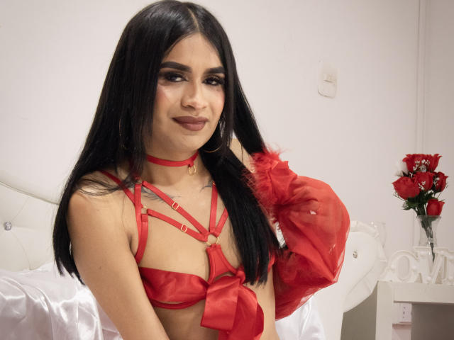 SilvanaCastro with long black hair, red strappy lingerie, puffy sleeves, and a red choker, smiling, showing small tits, in a white bedroom with red roses.