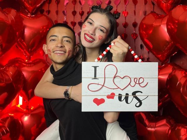 CrimsonSecretts, a cute woman with heart makeup, black cat ears, and a beaded necklace, hugs a smiling man with a black shirt, surrounded by red heart balloons.