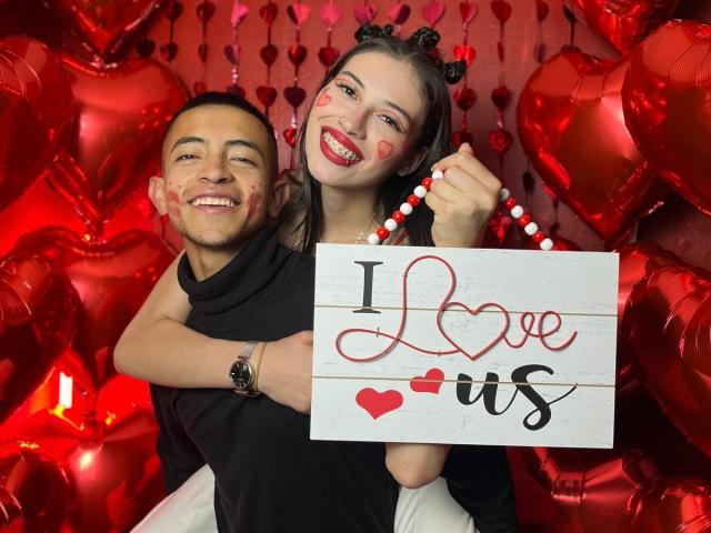 CrimsonSecretts with heart-shaped face paint, holding beads, stands in front of a smiling man, both surrounded by shiny red heart balloons. CrimsonSecretts has black cat ears, black sleeve.