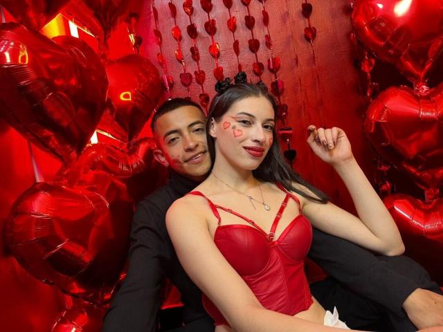 CrimsonSecretts with medium tits in a red leather bustier, face painted with a heart, sitting in front of a guy with short black hair, black shirt, surrounded by red heart balloons.