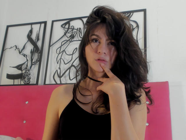 KariCarter has wavy, dark brown hair and is wearing a black choker and a black sleeveless top. She's holding her right index finger to her slightly parted lips. Her expression is neutral, with slightly pouty lips. The background has three black-and-white abstract framed art pieces and a pink, tufted headboard. The image focuses on her upper body and face. The lighting is even, highlighting her facial features and hair texture. Her skin tone is light. She has a slender build.