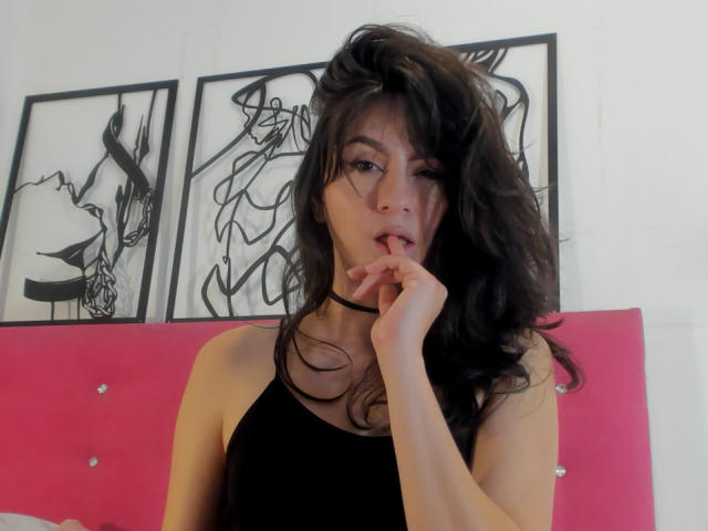 KariCarter has long, wavy black hair and a slightly messy look. She's wearing a black tank top and a black choker. Her right hand is near her mouth with her finger slightly touching her lips. She has a light skin tone and is looking directly at the camera with a slightly open mouth. Behind her is a pink, tufted headboard and three abstract black and white framed artworks on a white wall. The image is a photograph.