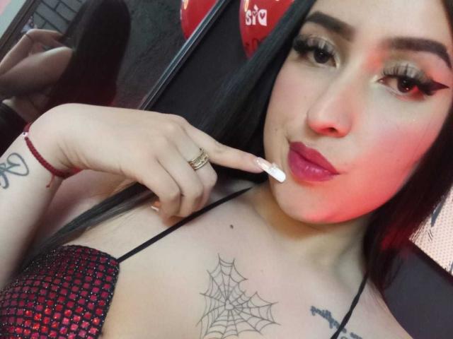 Chloey69 met lang zwart haar, rode lippenstift en eyeliner, houdt een witte stok tegen haar lip, toont een spinnenweb-tatoeage op haar borst, draagt een rode en zwarte top..