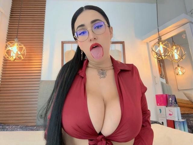 ScarlethSwann with big tits, tongue out, wearing glasses, red blouse, black hair in ponytail, necklace, and hoop earrings, in a bright room with hanging lights.