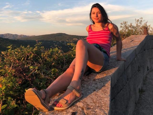 JainFresh-hot with medium tits, pink striped tank top, denim shorts, and sandals, sitting on a stone wall, legs crossed, tattoo on right thigh, sunset in background.