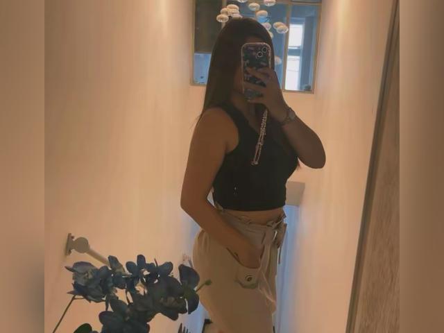 LinaFoxyHot with long dark hair, black top, and beige pants, holding phone, mirror selfie, showing ass, small tits, hands in pockets, blue plant in foreground.