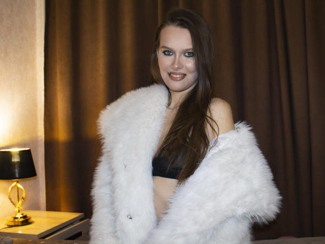 AyumiKirk with long brown hair, smiling, wearing a black bra and fluffy white coat, standing by a lamp, brown curtains in the background. Boobs visible under the coat.