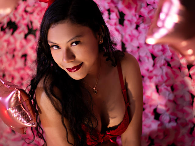 IvyMontaner with long black hair, red lingerie, and red lipstick, holding a pink balloon, smiling at the camera, showing her tits, pink flower background.