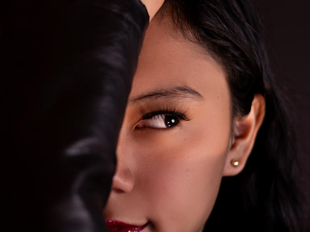 IvyMontaner's face with brown eyes, black eyeliner, red lipstick, and gold earrings, partially obscured by a black leather arm. Her skin is smooth and she has dark hair.