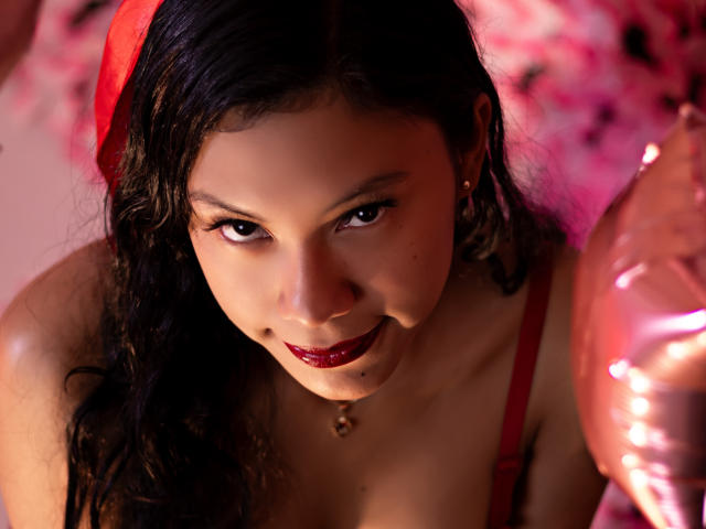 IvyMontaner with dark, wavy hair, red lipstick, and a red headscarf, looking up with a slight smile, wearing a red top, and a small necklace with a heart pendant.