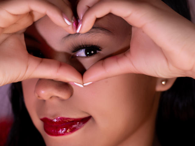 IvyMontaner's hands form a heart shape over her face, showing her brown eyes, glossy red lipstick, and manicured white nails with pink tips. She has a small beauty mark.