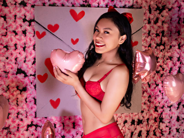 IvyMontaner with long black hair, red bra, and matching panties, holding pink heart-shaped balloon, smiling, pink and red decor, pink flowers, red hearts, and pink balloons around.