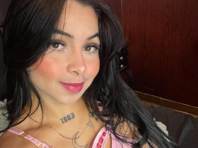 MistressAsshley with long black hair, pink lipstick, and a 1963 tattoo on her neck, wearing a pink bra, smiling at the camera. Boobs visible.